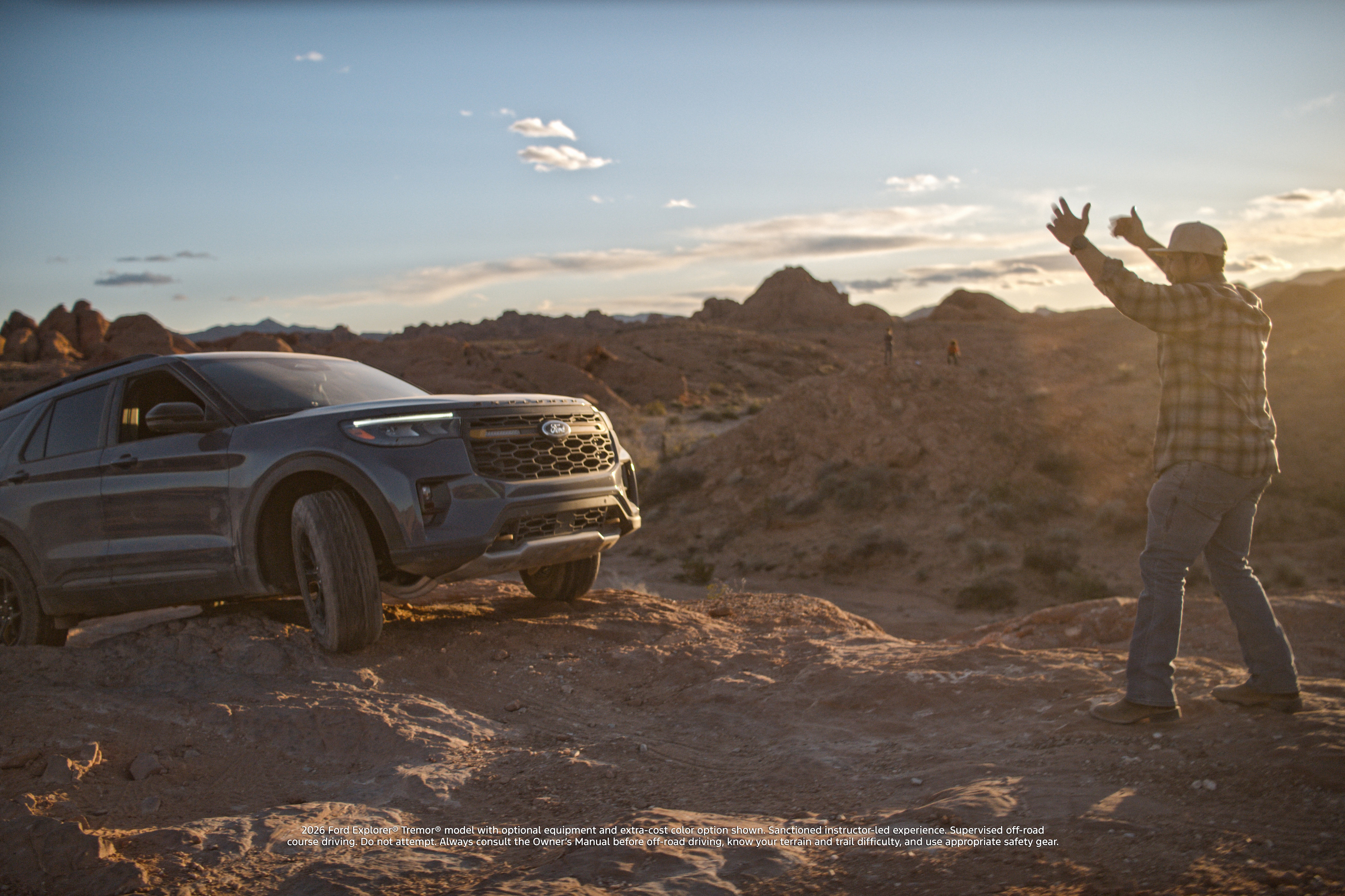 A man stands in front of a Ford Explorer on an off-road trail, guiding the driver with his hands up.